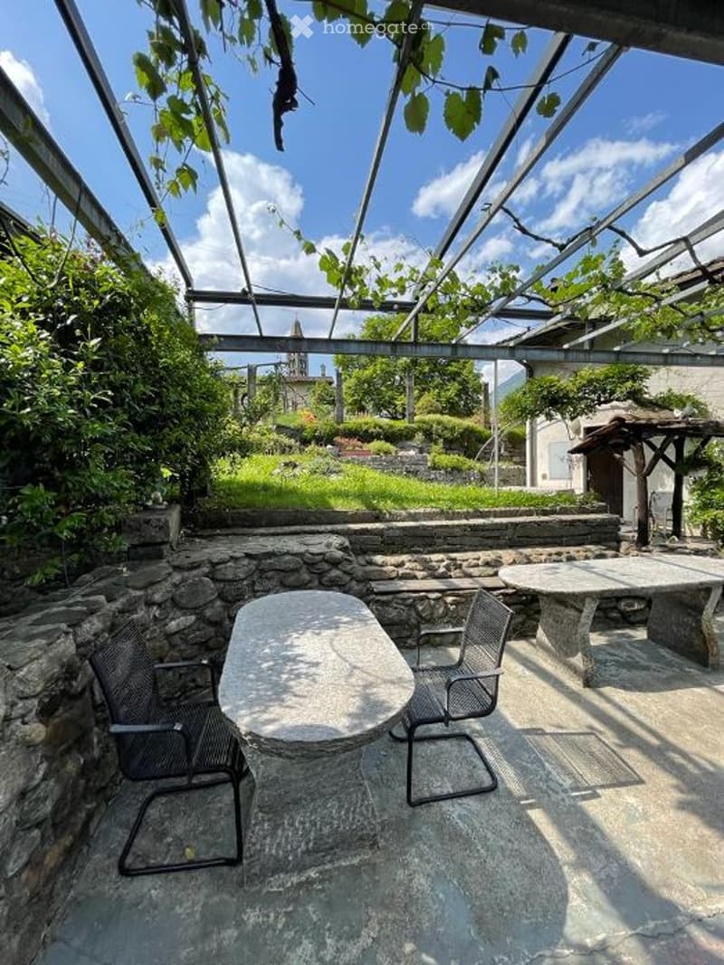 Outdoor seating area with stone walls, metal pergola, tables, chairs, and a view of a grassy hill and a building.