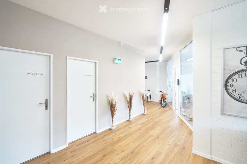 Empty hallway with two doors, wooden floor, white walls, three vases with dried plants, clock on the wall, emergency exit sign