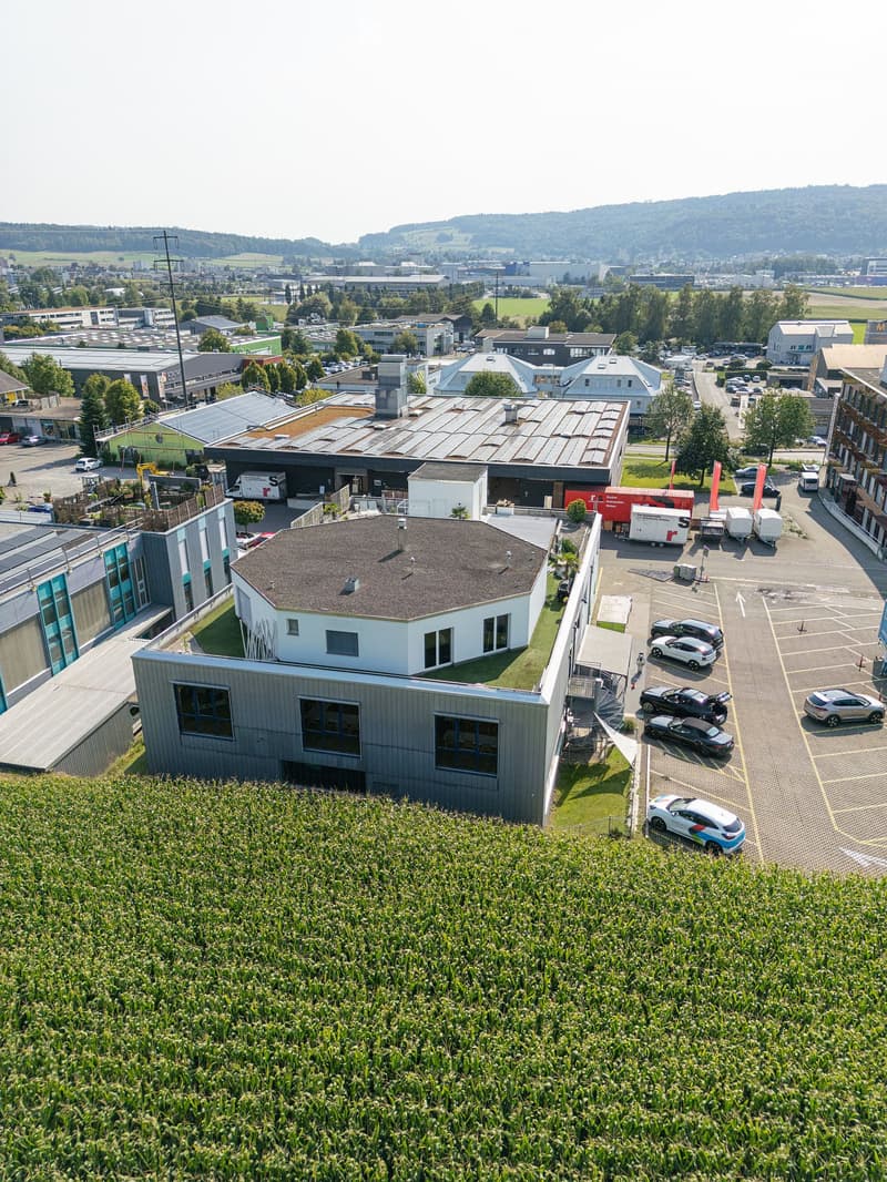 Modern building with a circular roof, surrounded by a parking lot, multiple buildings in the background, and a green field.
