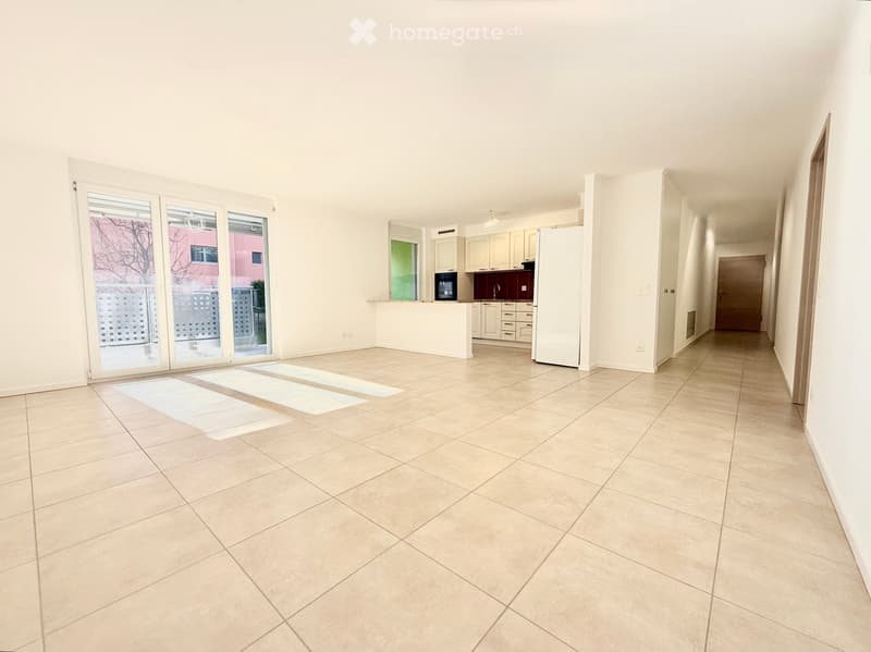 Empty room with tiled floor, sliding glass doors leading to a balcony, white walls, and a kitchen area with white cabinets and a refrigerator.