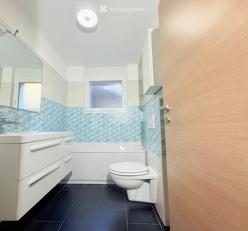 Bathroom with white bathtub, white toilet, white sink, blue tiles, white cabinets, mirror, and a window