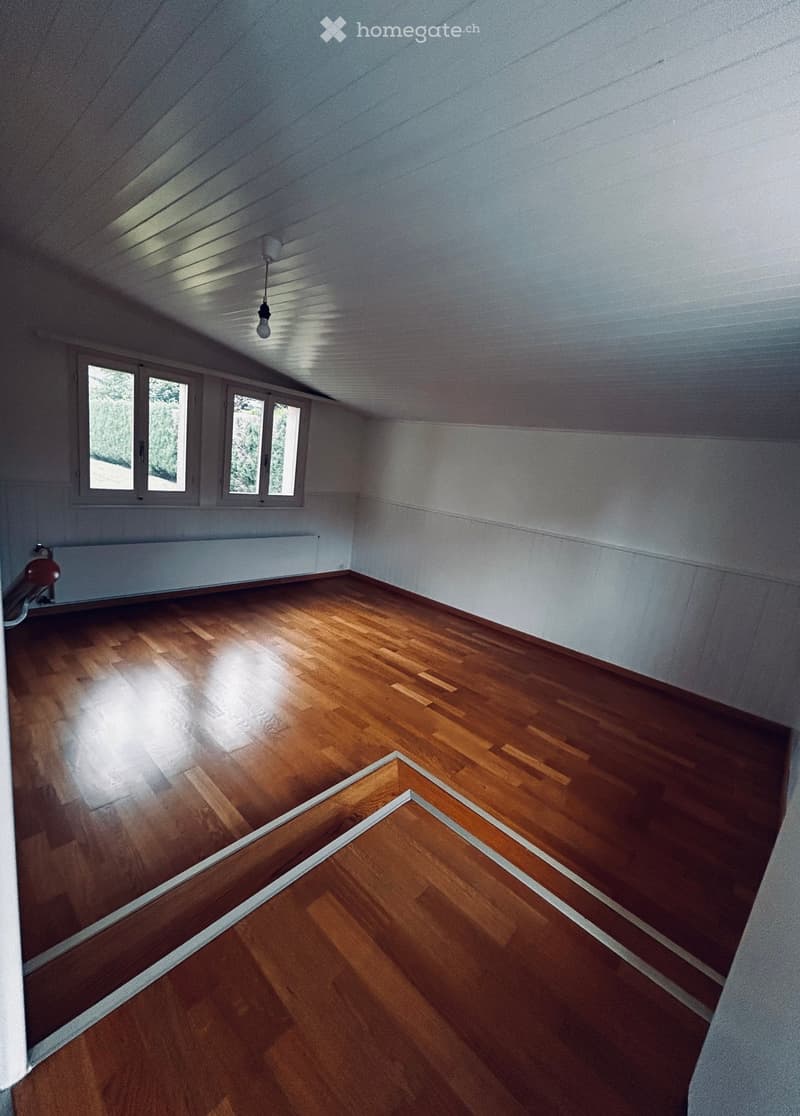 Empty attic room with wooden flooring, white walls, and large windows allowing natural light. The ceiling has exposed wooden beams and a single hanging light fixture.