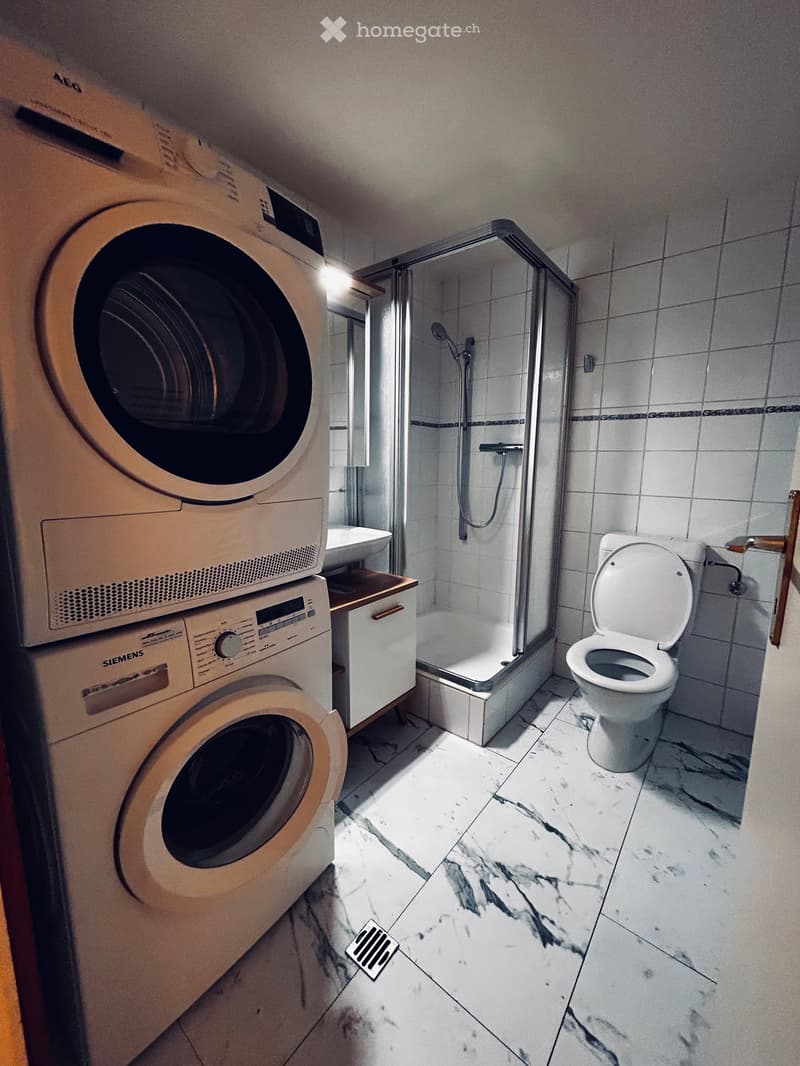 Modern bathroom with marble flooring, a toilet, a shower, a sink, and stacked washing machine and dryer units.