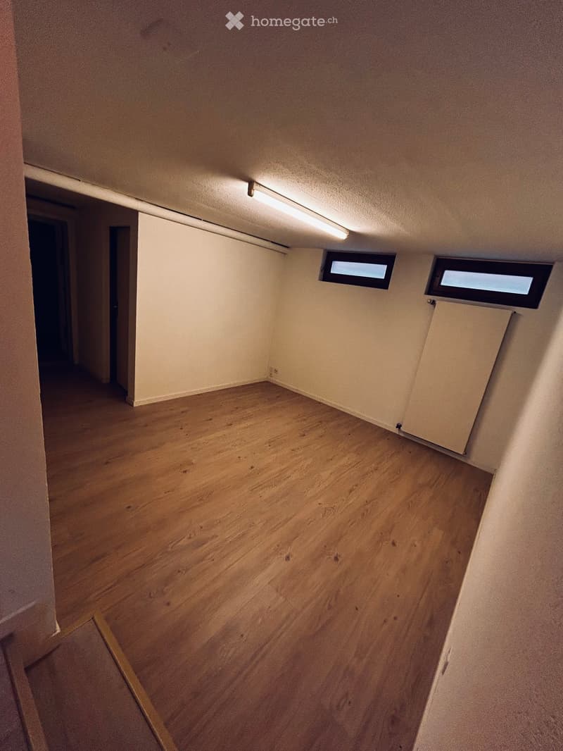 Empty room with wooden flooring, white walls, and two small windows near the ceiling. The room has a door leading to another space.