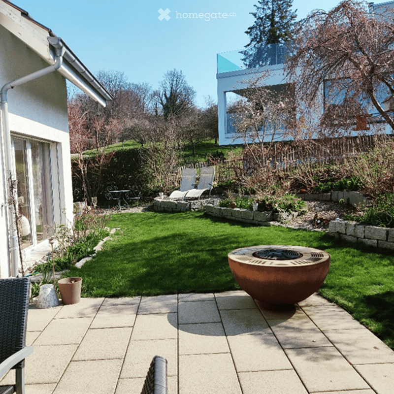Patio with a fire pit, chairs, and a table, surrounded by a well-maintained lawn and garden with various plants and trees.