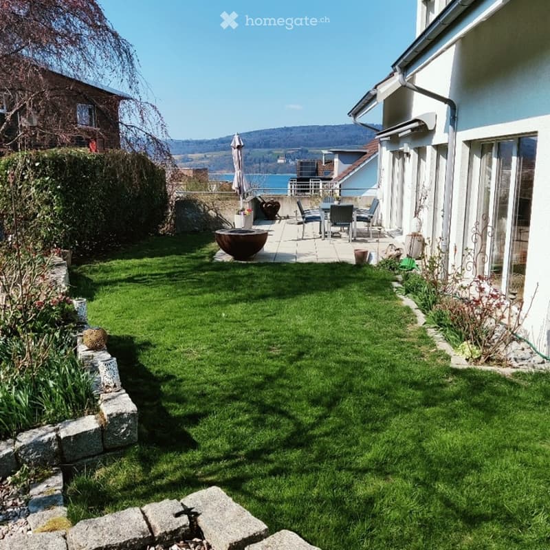 well-maintained garden with green lawn, stone border, outdoor seating area with table and chairs, potted plants, and a view of the lake