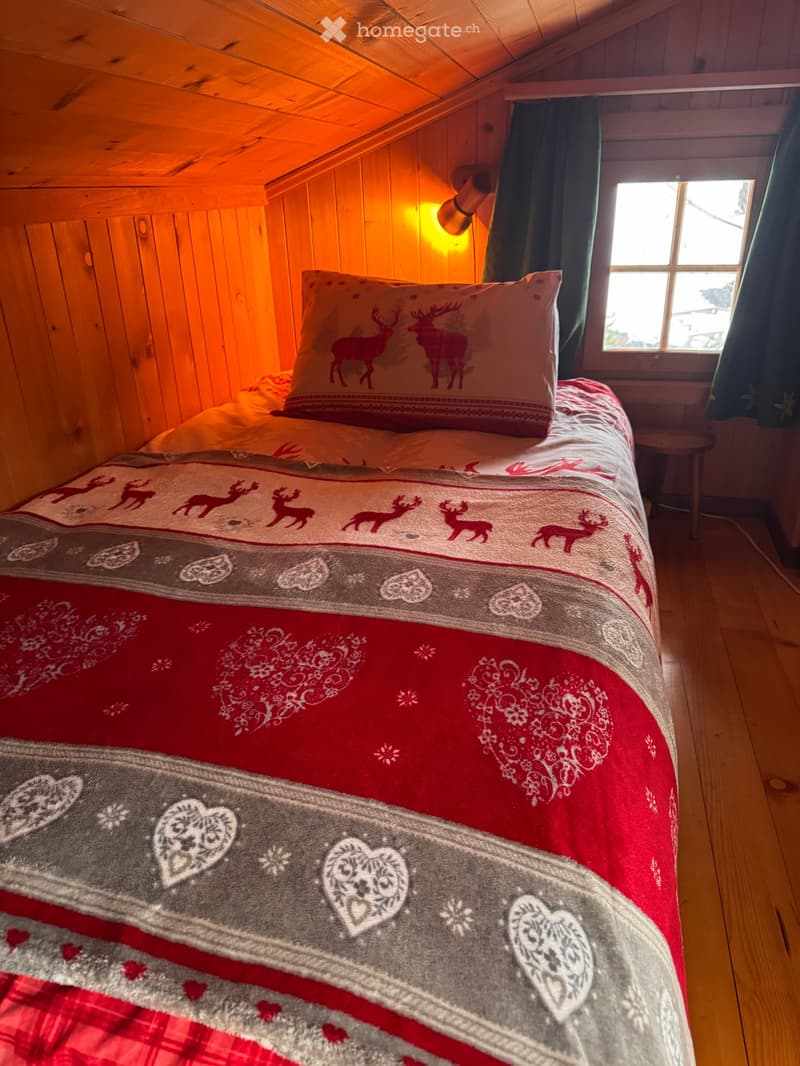 Bedroom with wooden walls, bed with red and gray bedspread, wooden floor, window with green curtains, wall lamp, small wooden stool