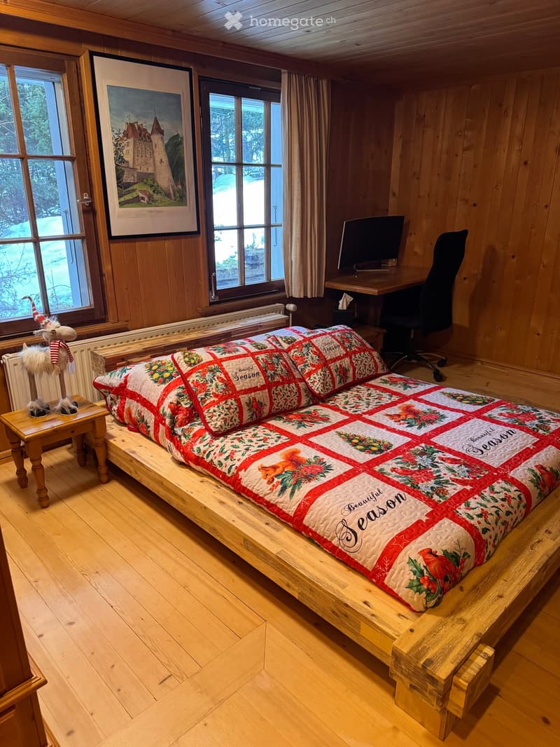 bedroom with wooden floor, bed with quilt, desk with computer, chair, radiator, window, picture frame, wooden side table with reindeer statue
