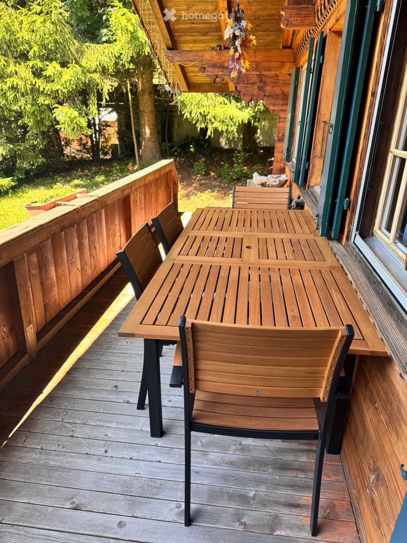 Wooden table and chairs on a wooden deck, surrounded by a wooden railing, with a view of the grassy area and trees