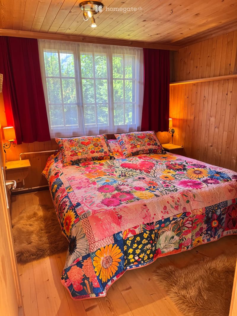 Bedroom with wooden walls, bed with floral quilt, two nightstands with lamps, rug, large window with white curtains, red curtains