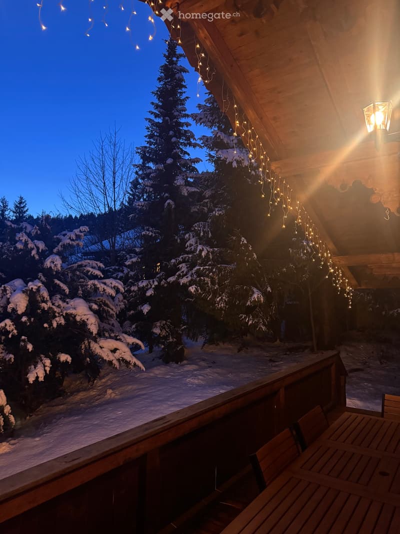 wooden balcony with railing, outdoor table and chairs, string lights, snowy trees, snowy ground, blue sky