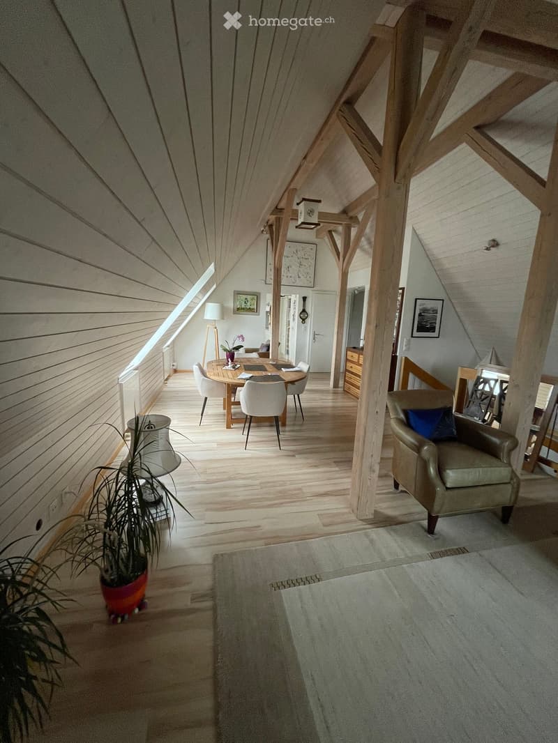 A cozy attic room with wooden beams, wooden floor, a round dining table, and a wooden cabinet. There is a couch with a pillow, a potted plant, and a lamp.