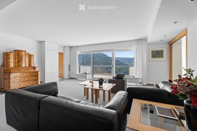Spacious living room with large windows offering a scenic view of the surrounding mountains and forests. The room features a black leather sofa, a wooden coffee table, and a wooden dresser. The overall decor has a modern and minimalist style.