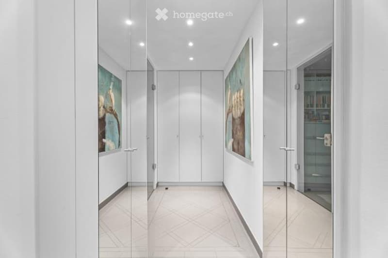 Bright, modern hallway with white walls, recessed lighting, and large framed artwork on the walls. The floor is tiled with a geometric pattern. There appears to be a glass door or entryway visible at the end of the hallway.