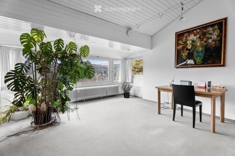 Spacious living room with large windows, white walls, hardwood floors, and a large potted plant. There is a desk with a chair and a framed artwork on the wall.