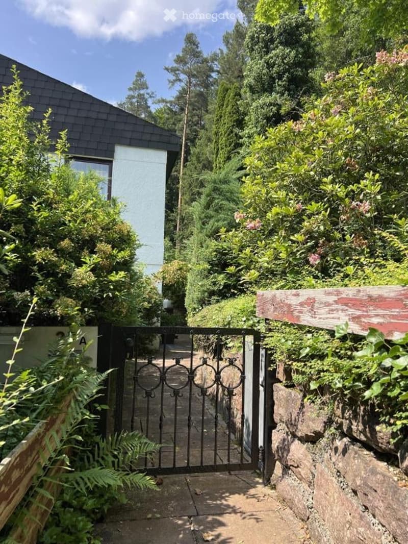 2-story house with black roof tiles, surrounded by lush greenery and vegetation, including trees, shrubs, and ferns. There is a metal gate leading to a path or walkway.