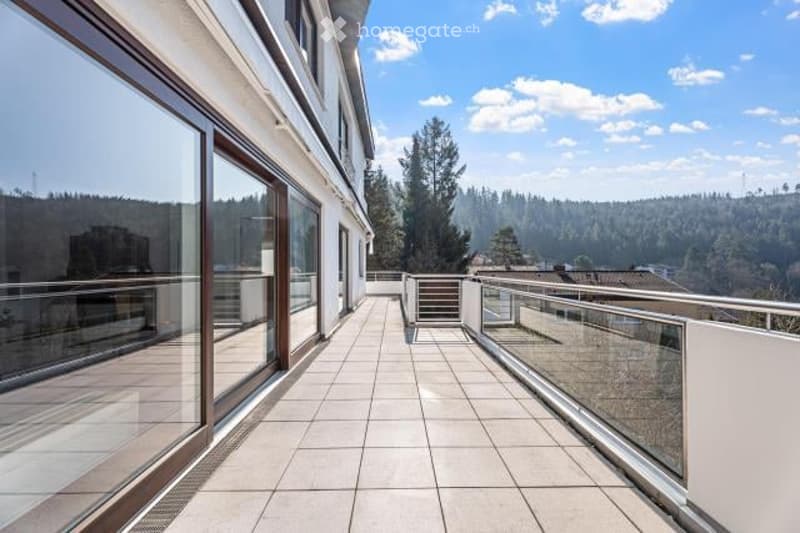Large modern house with a spacious balcony or terrace overlooking a forested area. The balcony has glass railings and sliding glass doors leading to the interior. The exterior of the house appears to be made of light-colored materials, and the surrounding