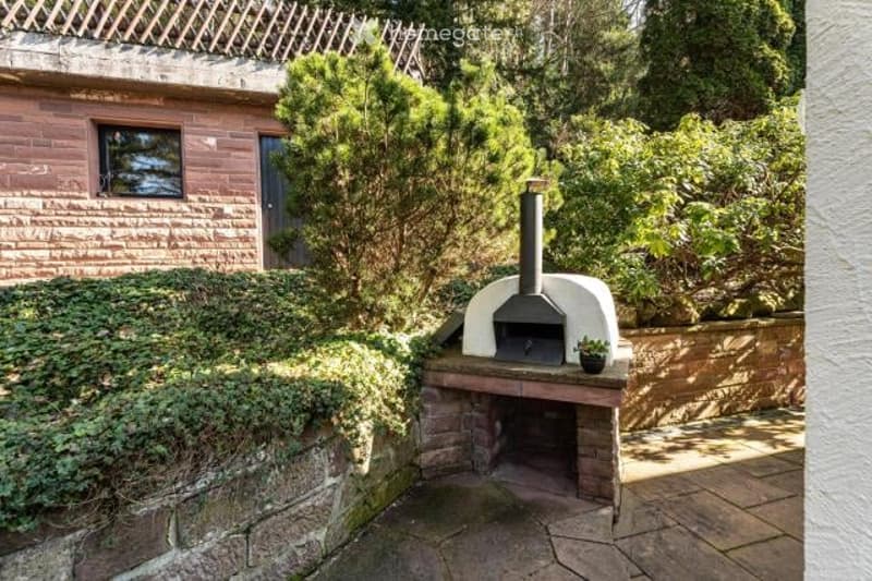 Outdoor patio area with a brick and stone pizza oven or fireplace, surrounded by greenery and vegetation.