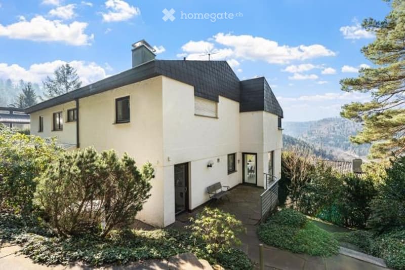 Two-story house with a white exterior, black roof, and large windows. The house is surrounded by greenery and has a balcony or terrace on the upper level.