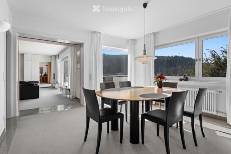 Spacious dining room with large windows offering a scenic view, wooden dining table with black leather chairs, pendant lighting fixture, open floor plan leading to other rooms
