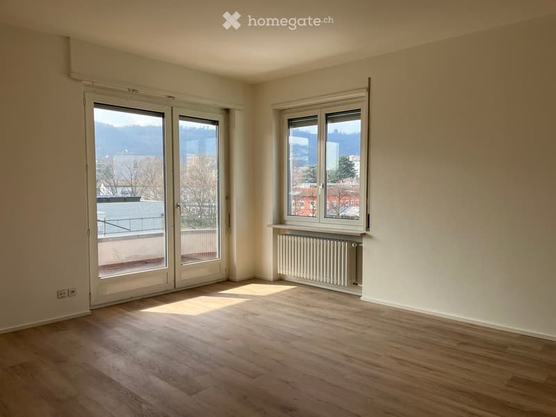 Empty room with wooden flooring, large windows, radiator, and a balcony door leading to an outdoor area.