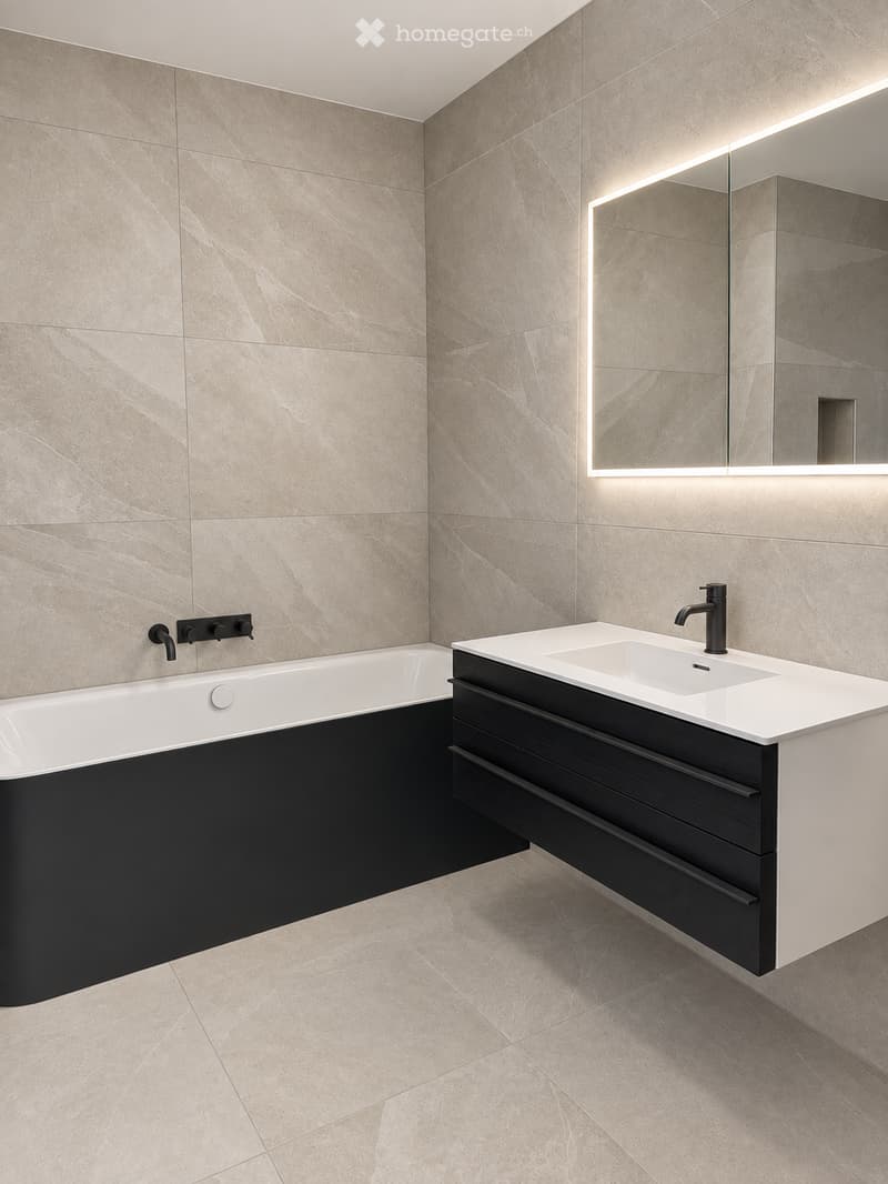 Modern bathroom with a white bathtub, black vanity, and a sleek sink with a modern faucet. The walls and floor are covered in light beige marble tiles, and a large mirror with integrated lighting is mounted above the sink.