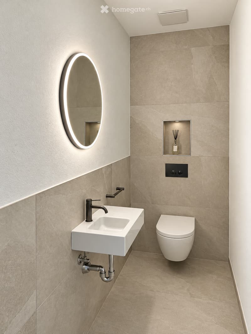 Modern bathroom with a white sink, sleek faucet, toilet, illuminated oval mirror, and a small shelf holding a bottle of perfume.