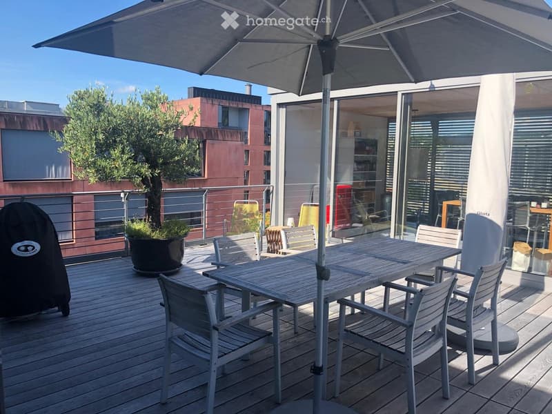Wooden deck with table and chairs, large umbrella, potted plants, metal railings, distant buildings