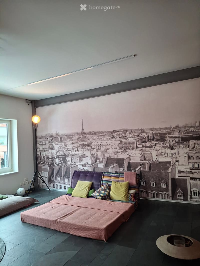 A room with a large mural of a cityscape, a bed with colorful pillows, a lamp, and a round table.