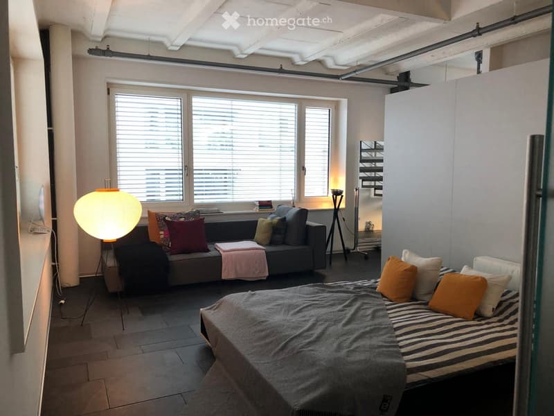 A room with a bed, couch, lamp, and windows with blinds.