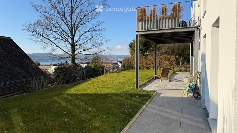 A garden with a lawn, a metal pergola, a wooden chair, a hose reel, and a view of the city and lake.