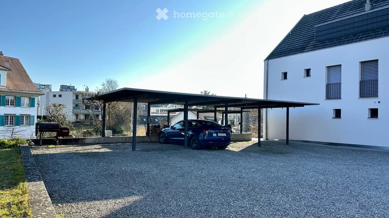 Car parked under a carport, white building, gravel driveway, solar panels on the roof