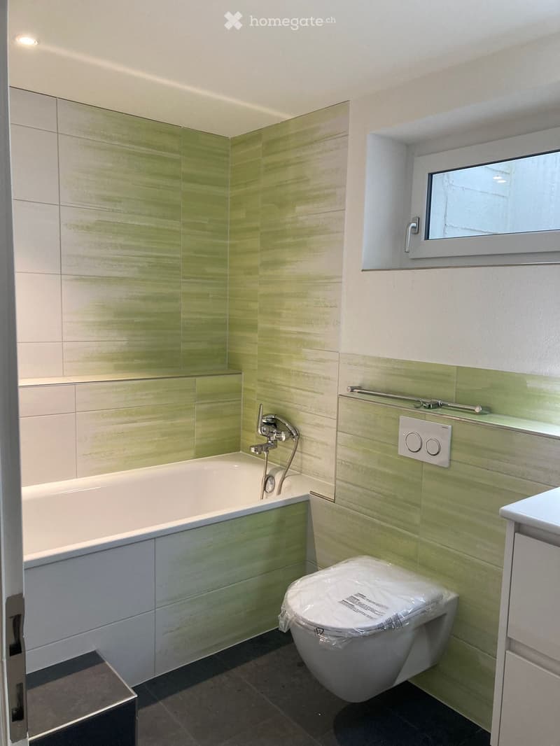 Bathroom with green and white tiles, bathtub, toilet, sink, and a window.