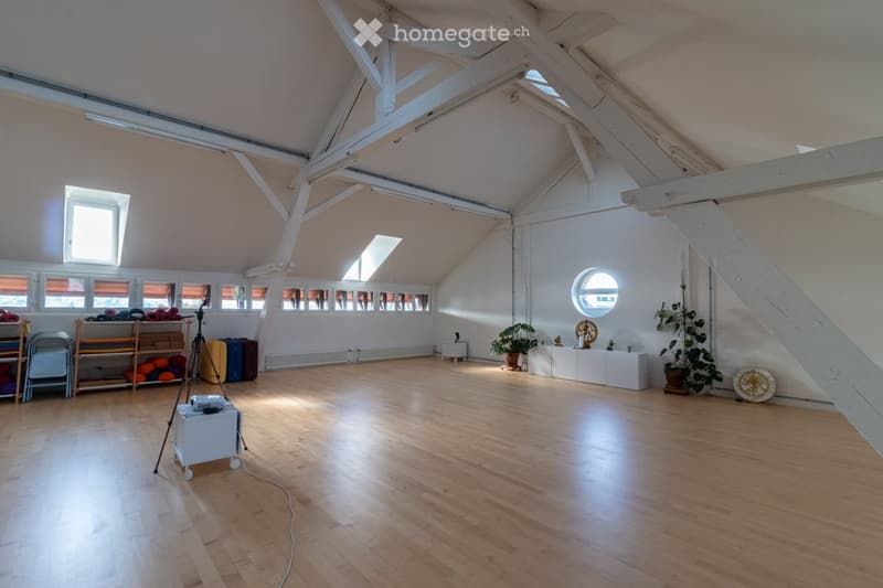 large open space, wooden floor, white walls, wooden beams, windows, plants, tripod with projector