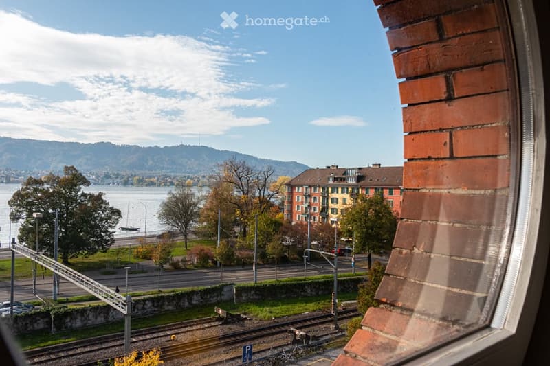 View from a window of a building overlooking a lake, mountains, and a road with a train track.
