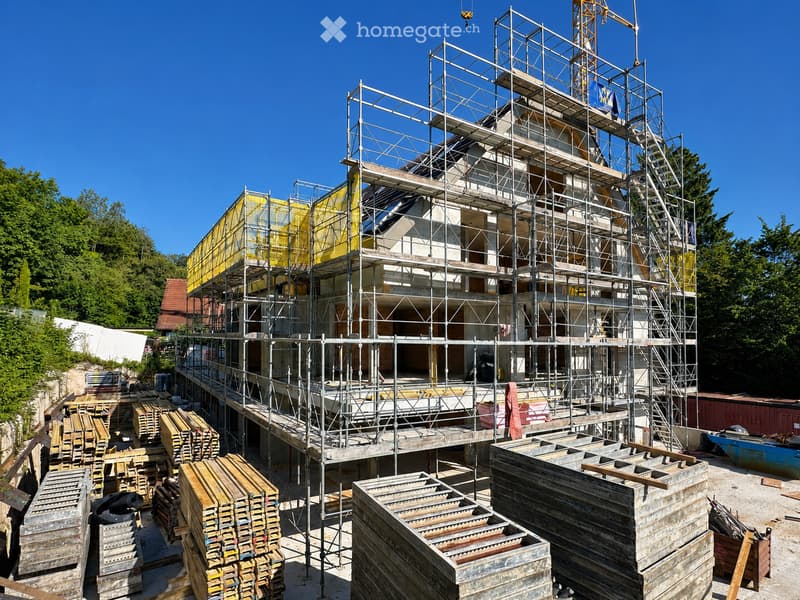 A multi-story building under construction, covered in scaffolding with exposed steel frames and partially covered with yellow netting. Construction materials and tools are scattered around the base.