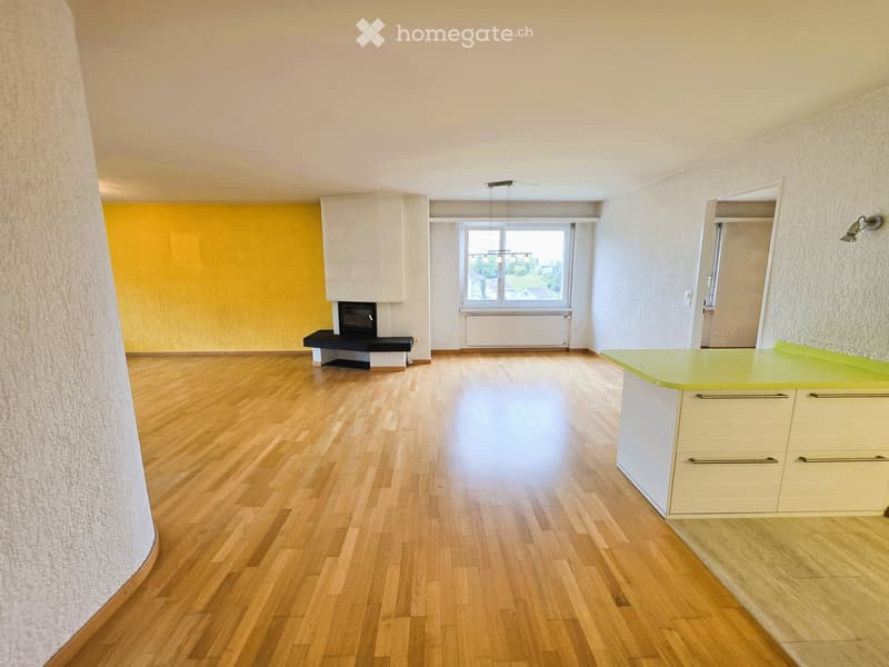 Bright, open room with wooden flooring, a small kitchen area with cabinets, a fireplace, and a window offering a view of the outdoors.