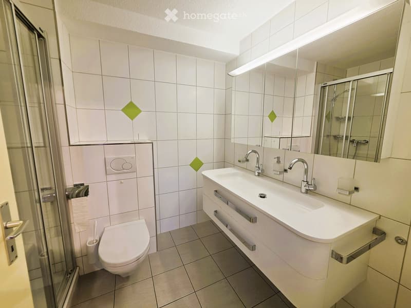 Modern bathroom with white tiles, a double sink, a toilet, a shower area, and a large mirror with built-in lighting.