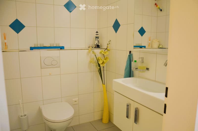 white tiles, white toilet, white sink, yellow vase with flowers, blue diamond tiles, white toilet paper holder, soap dispenser, towel rack