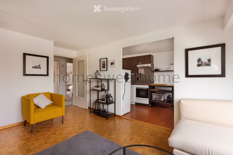 living room with wooden floor, white walls, yellow chair, white sofa, glass table, open kitchen with appliances, framed pictures on the wall
