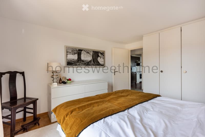 white walls, wooden floor, bed with white sheets and brown blanket, white dresser, lamp, picture frame, wooden chair