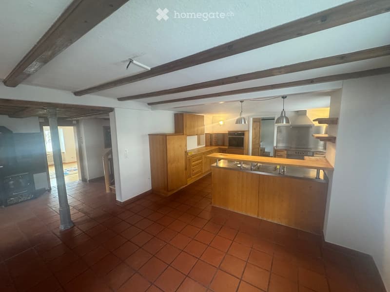 Open kitchen with wooden cabinets, red tiled floor, wooden beams, central island, hanging lamps, stove, and a fireplace.