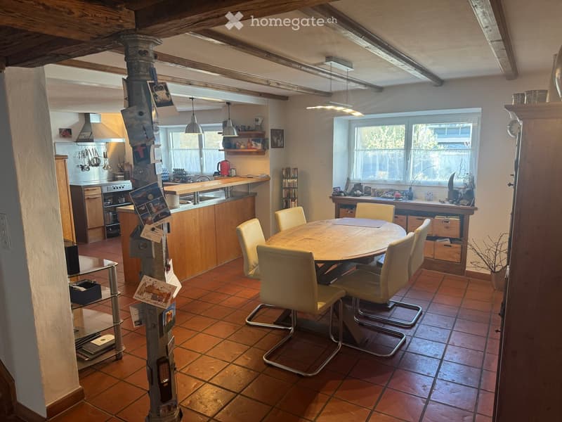 Dining room with wooden table and chairs, kitchen area with counter, cabinets, and appliances, wooden beams on ceiling, tiled floor, and large window.