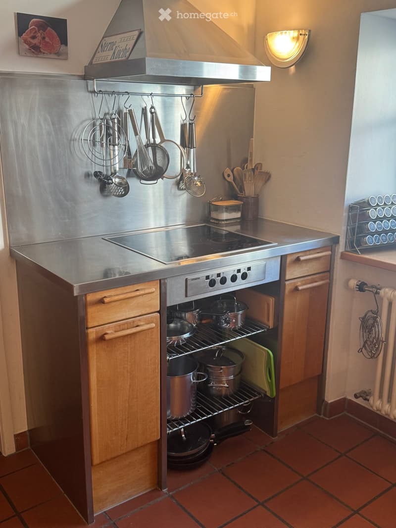 Stainless steel countertop, wooden cabinets, gas stove, stainless steel hood, hanging utensils, wooden spoons, spice rack, radiator