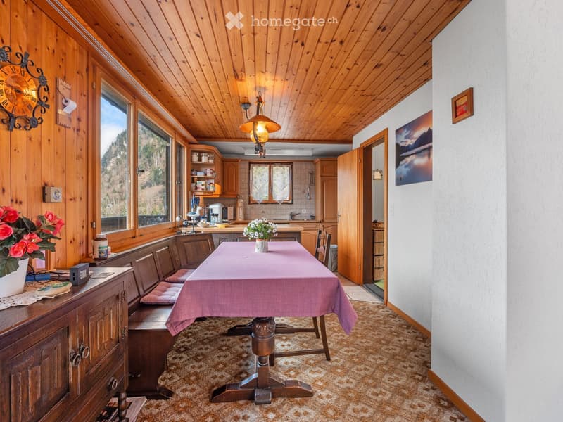 Dining room with wooden walls, wooden table, bench seating, flower vase, kitchen area, wooden cabinets, picture frames, carpeted floor