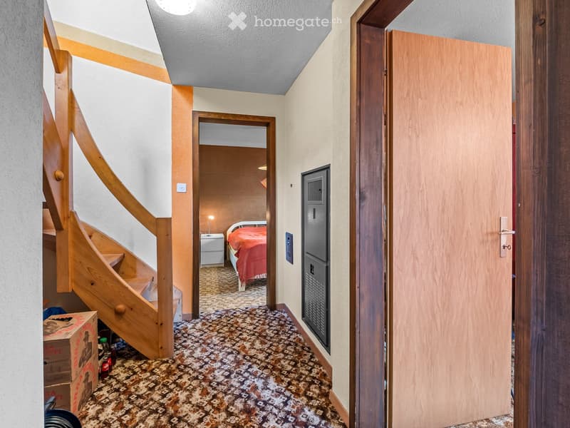 A hallway with a wooden staircase, a bedroom with a bed and nightstand, and a closed door.