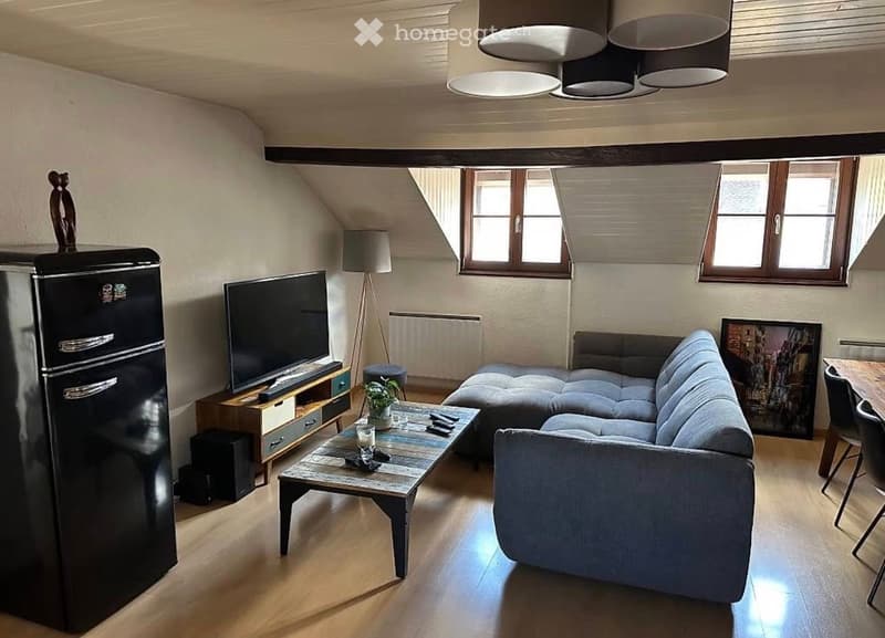 Living room with wooden floor, gray sofa, wooden coffee table, flat screen TV, wooden entertainment center, black refrigerator, wooden dining table, black chairs, lamps, picture frame, radiator, wooden ceiling beams
