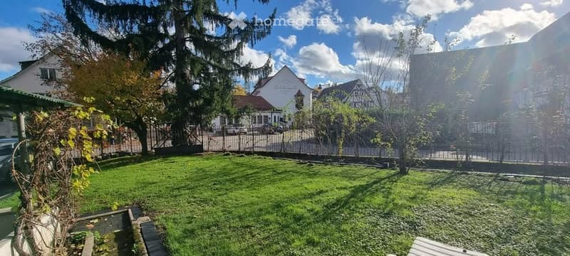 green lawn, trees, shrubs, black metal fence, black metal gate, parking lot with parked cars