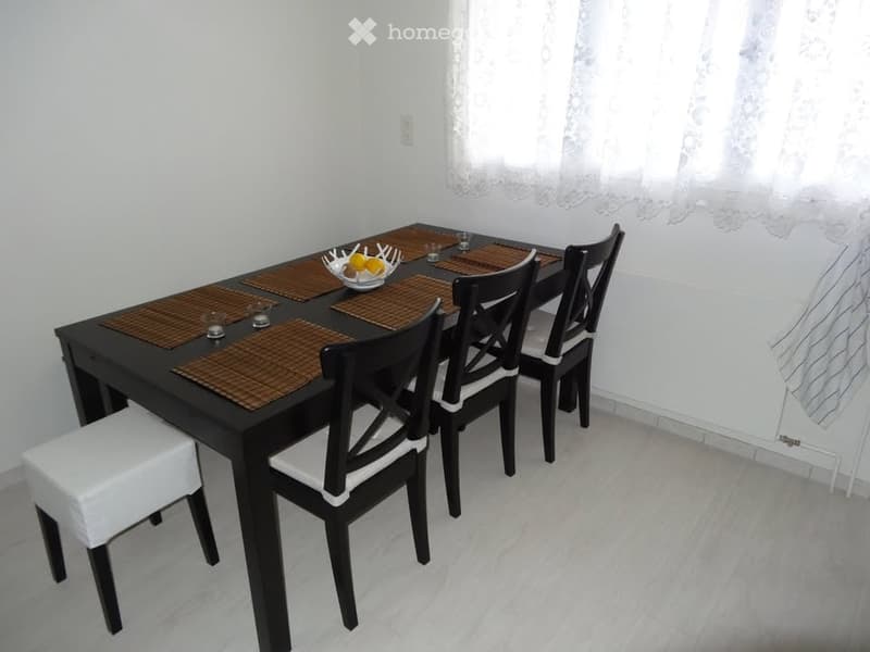 Dining room with a black dining table, four chairs, and a white bench. Centerpiece is a bowl of lemons. White curtains and radiator behind the table.