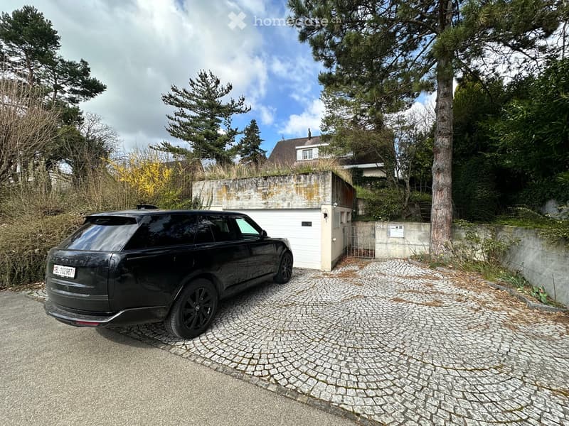 House with garage, black SUV parked in front, cobblestone driveway, trees and plants surrounding the area